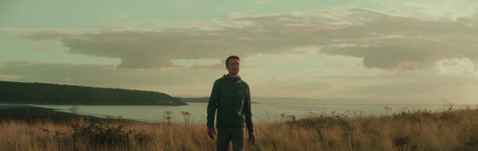 A person is walking through a grassy field towards a calm expansive lake with a forested shoreline and a cloudy sky in the background