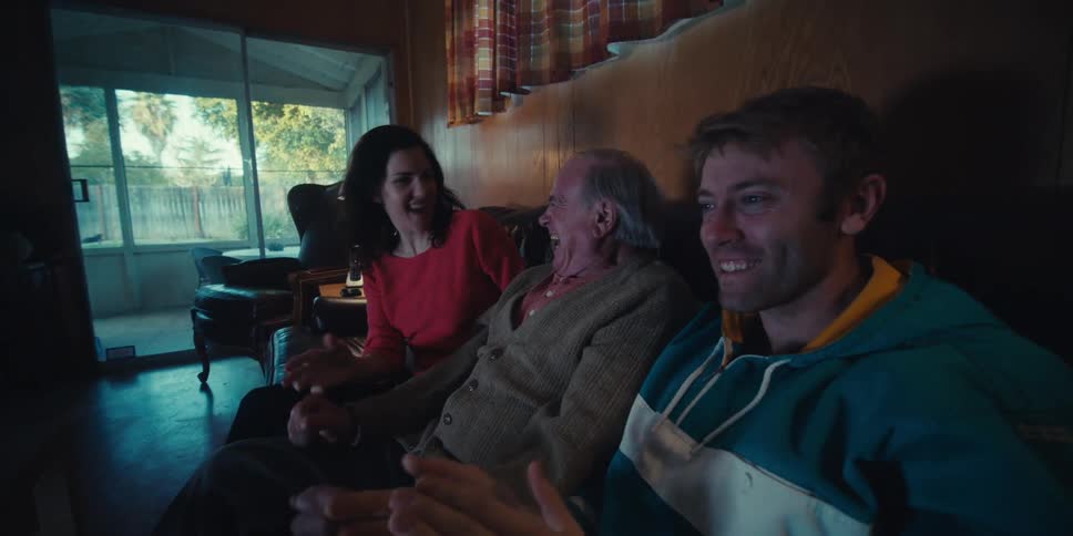 Three people a younger man and woman and an elderly man react to watching something on television sitting on the couch
