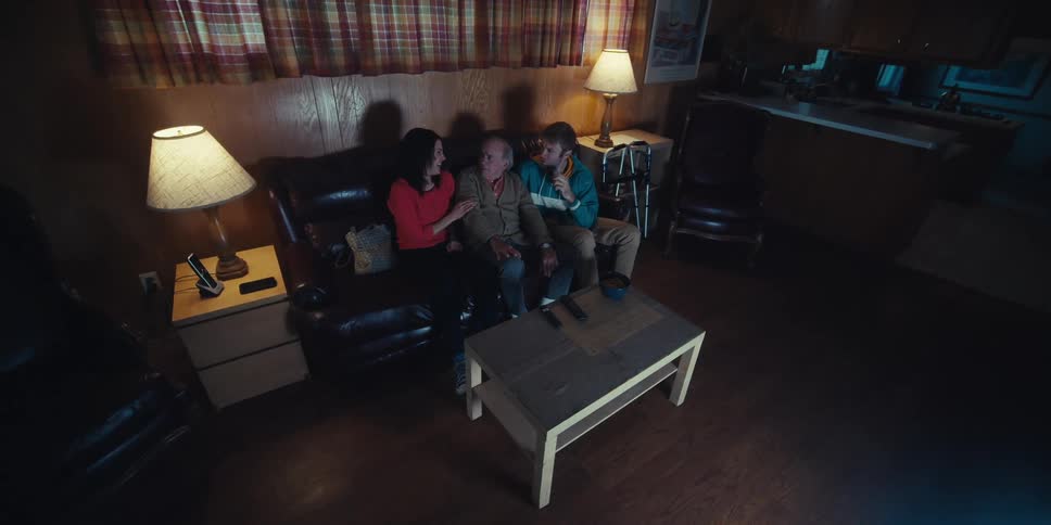 A family of three is sitting on a couch in a dimly lit room watching television together