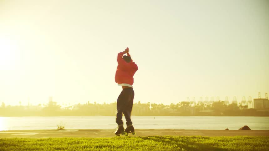 A man wearing a red sweater dances and spins on roller skates with arms raised near a waterfront at sunset