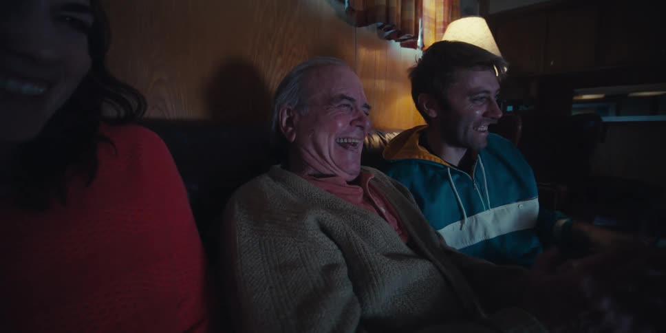 A family of three is sitting on a couch in a dimly lit room watching television together