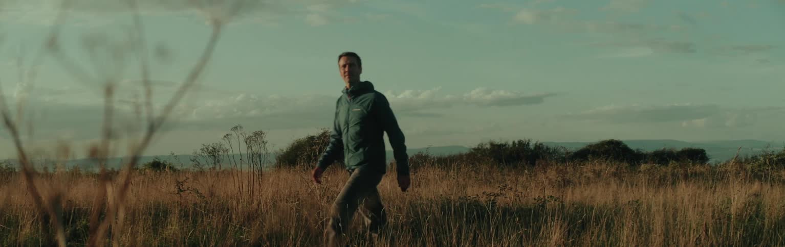 A person wearing a blue jacket is walking through a field of tall dry grass with bushes and a distant mountain range under a cloudy sky