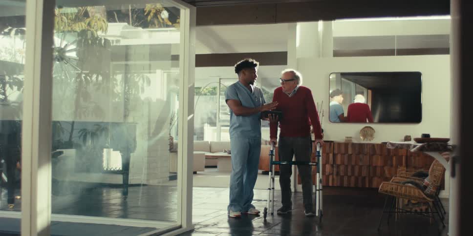 An elderly person using a walker is being assisted by a caregiver in a modern spacious living room with a grand piano and large windows
