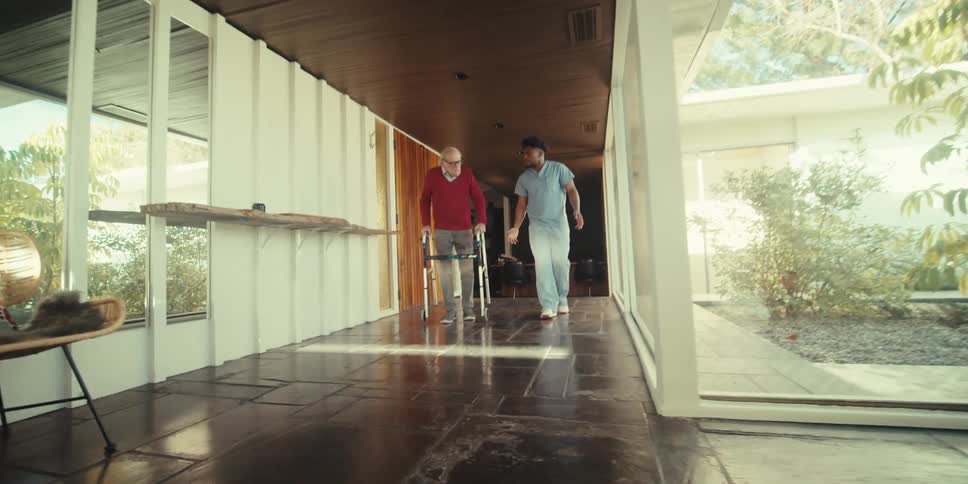 A healthcare worker is assisting a man using a walker down the hall