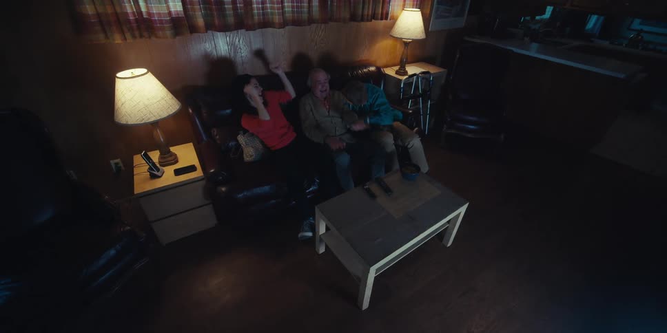 A family of three is sitting on a couch in a dimly lit room watching television together
