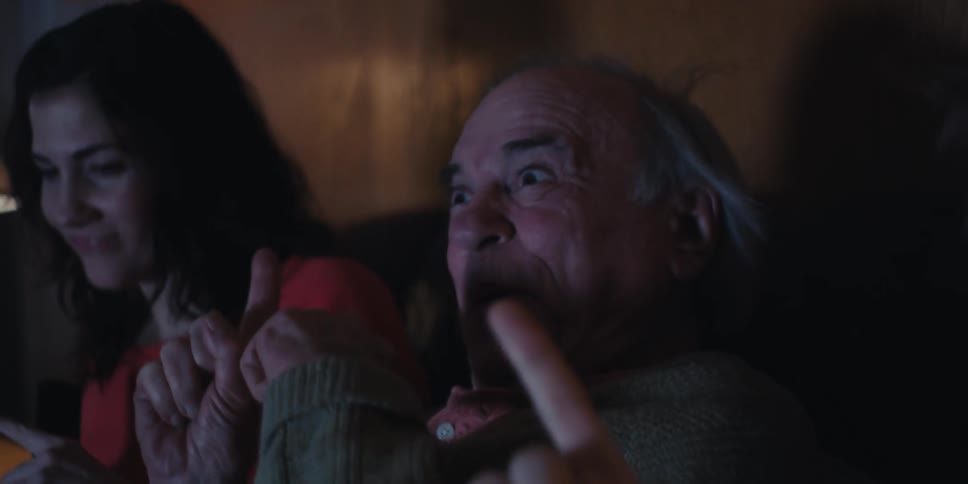 Elderly man sitting in the middle of two young men laughing as they watch together sitting on the living room couch