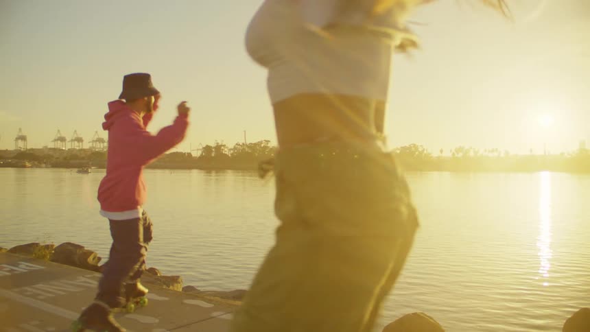 A couple of dancers perform energetic dance moves on roller skates near a waterfront at sunset