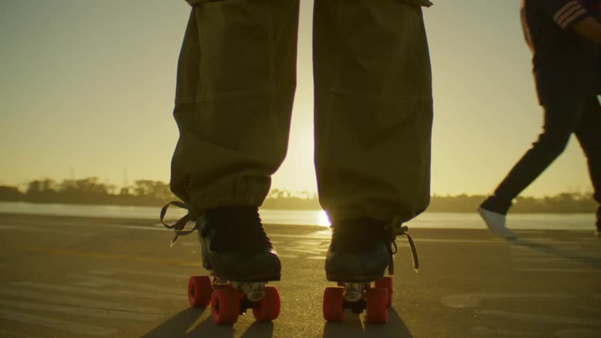 A person wearing roller skates stands waiting for others to pass before starting to dance near a body of water at sunset