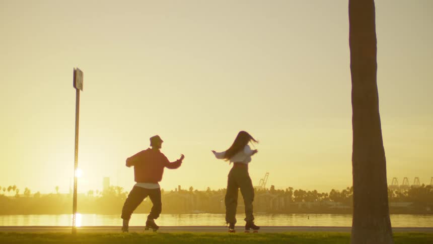 Woman and a man dancing on roller skates on the edge of a lake at sunset and the city in the background