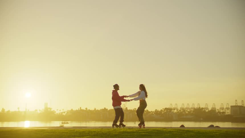 Two people on roller skates are dancing with their arms raised near a waterfront at sunset with an industrial skyline in the background