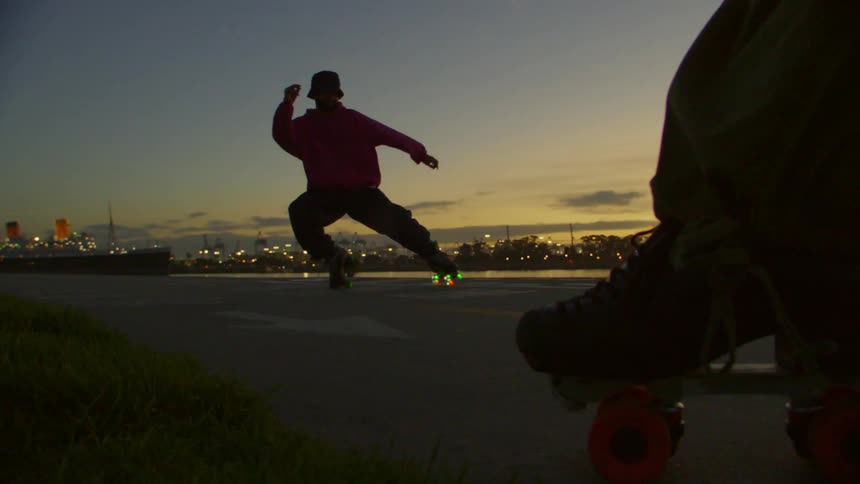 A person in a pink hoodie and black pants is roller skating on a paved surface at dusk with city lights and a waterfront visible in the background at dusk