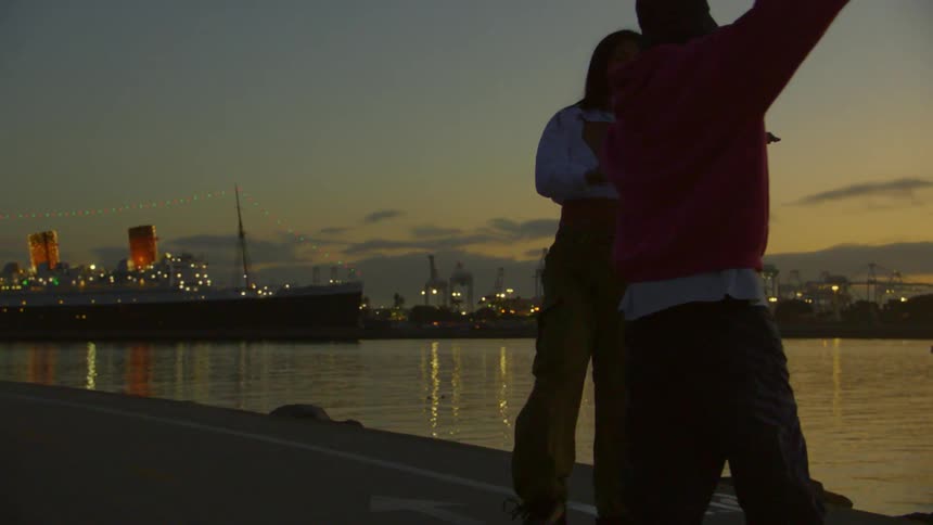 Two people dancing on roller skates at a waterfront during dusk while a large illuminated ship is visible in the background