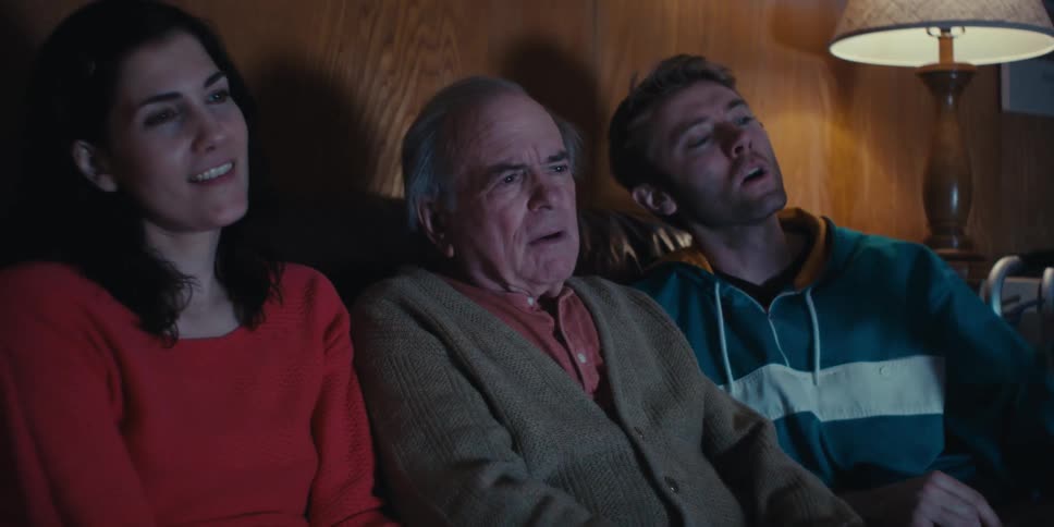 A family of three is sitting on a couch in a dimly lit room watching television together