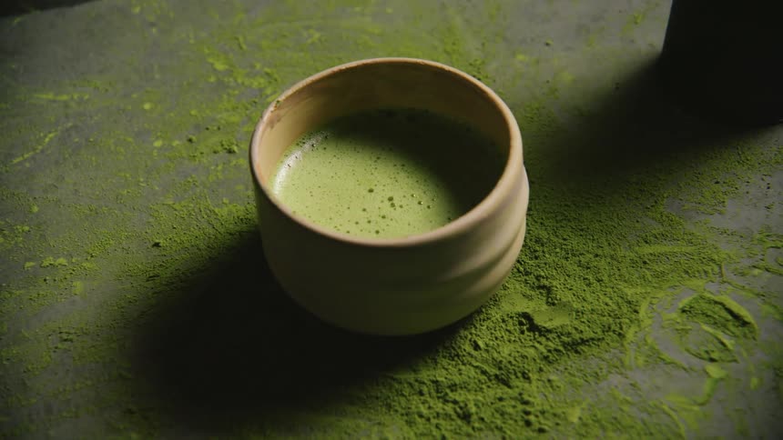 A and places a ceramic bowl filled with frothy green matcha tea on a surface dusted with matcha powder