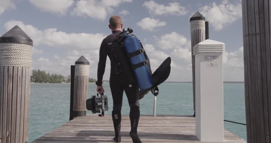 A scuba diver carrying gear is walking toward the end of a pier in daylight