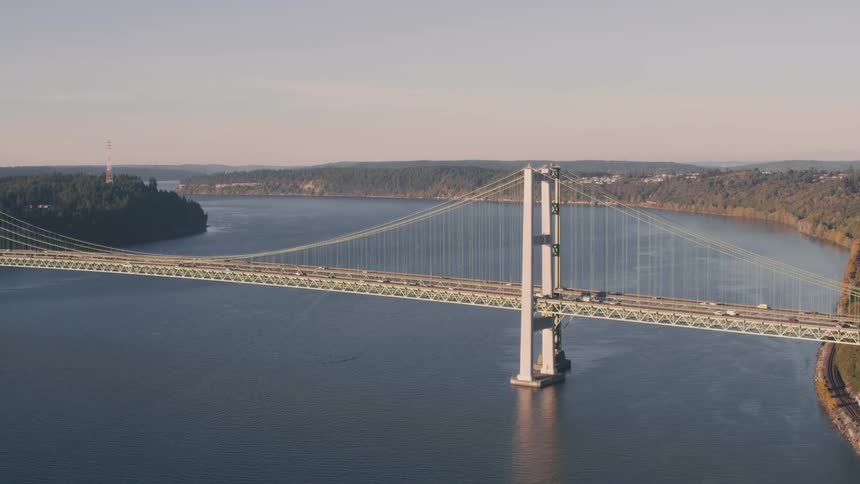 The tacoma narrows bridge spans a wide calm body of water connecting two forested shorelines under a clear sky
