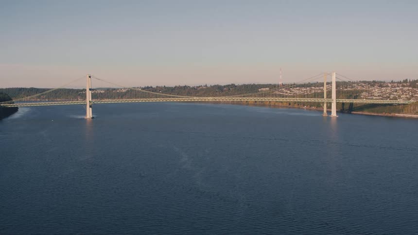 A suspension bridge spans across a wide river with an urban area on the right under a clear sky