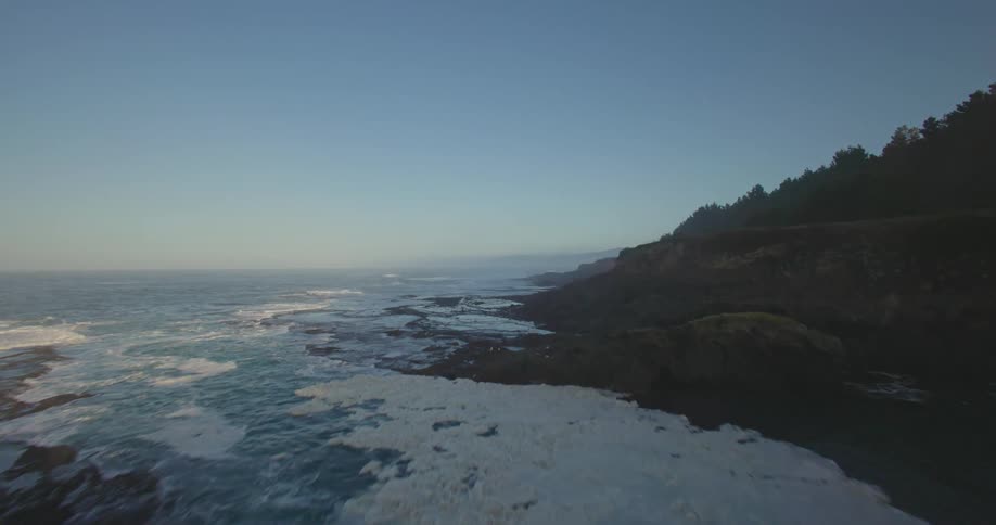 Vegetated cliffs stretch out over the blue ocean where waves gently break beneath a clear sunlit sky