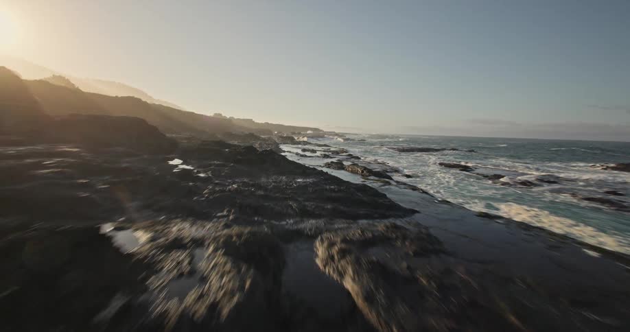 Golden sun sets over a rocky coastline where waves gently break against the wild shore