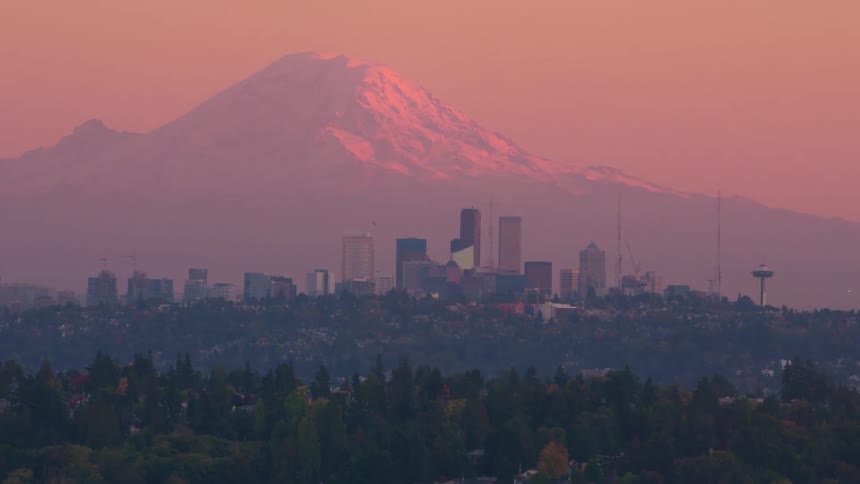 A majestic snowcapped mountain looms over a distant city skyline framed by a hazy reddishpink sky