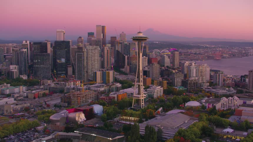Seattle lights up under a pink sky at dusk with the space needle standing out among skyscrapers and the bay in the background
