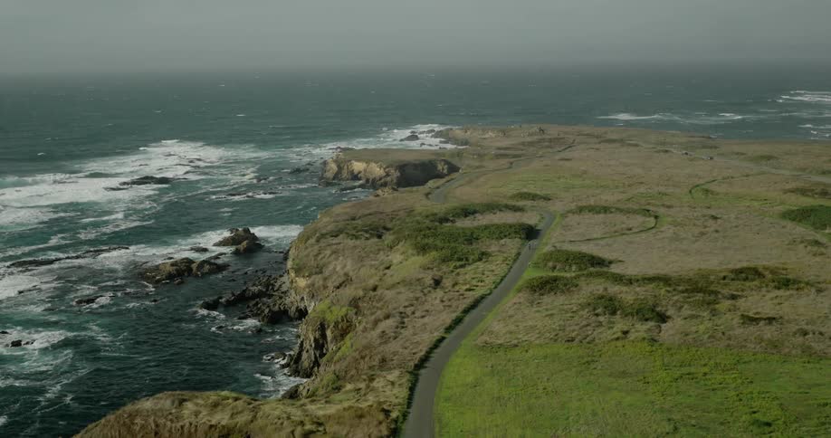 A rugged coastline with rocky cliffs and waves crashing against the shore alongside a winding road that traverses through a grassy sparsely vegetated landscape