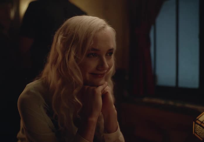A woman with long wavy blonde hair is sitting at a table in a dimly lit room with a window and a lamp visible in the background