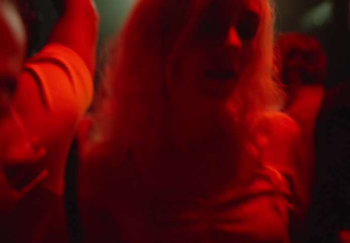 A blonde woman dances among a crowd of people in a dimly lit possibly smoky room with changing colored lights