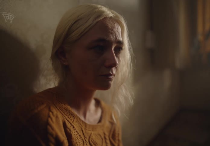 A woman with disheveled hair and smudged makeup sits on the floor against a worn wall wearing a knitted sweater with a sorrowful expression as dim light filters through a nearby window