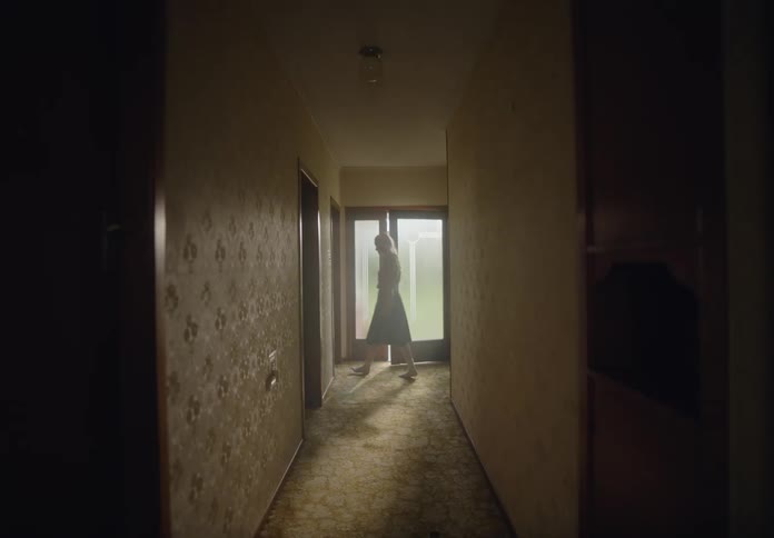 A woman is moving cardboard boxes at the end of a dimly lit hallway with light streaming in from a glass door behind them