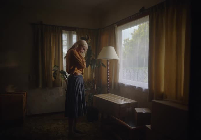 A woman is standing in a dimly lit room looking out of a window with yellow curtains next to a lamp and some furniture