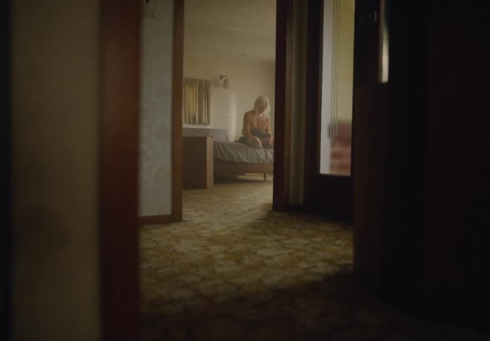 Blonde woman holds an object in her lap while sitting on the bed with a downcast expression