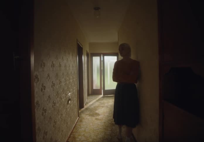 A woman walks through a dimly lit hallway carrying boxes with sunlight streaming in through a glass door at the end