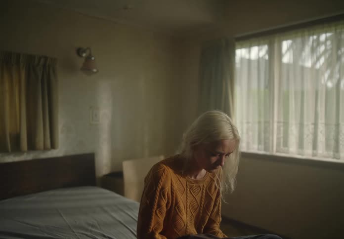 A sad blonde woman sits on the edge of a bed in a dimly lit room with a window and curtains
