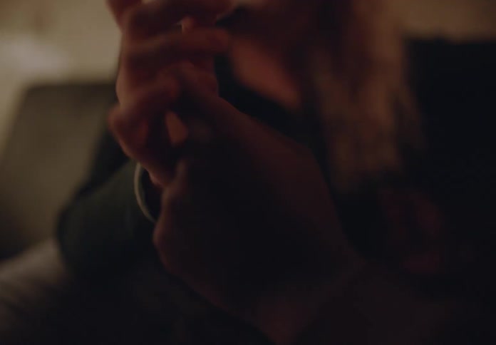 A young couple sits on a couch in a dimly lit room their hands interlocked as they gently caress each other