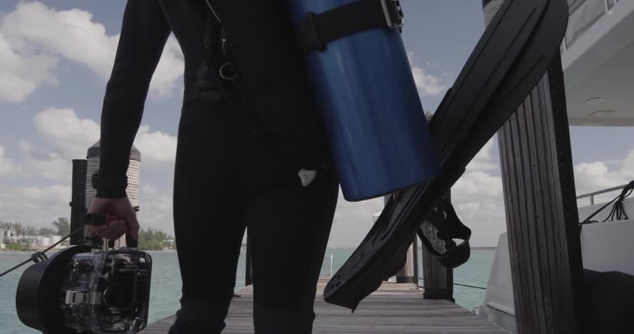 Scuba diver carrying his equipment and a camera towards the end of a pier on a partly cloudy day