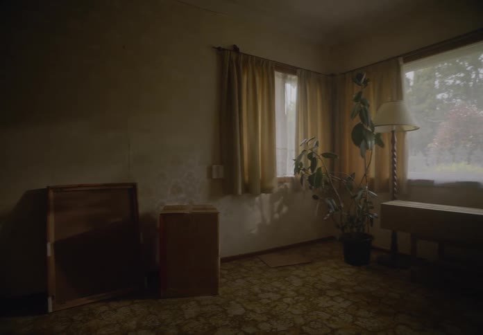 A person is sitting alone at a table in a dimly lit room with closed curtains and a window in the background