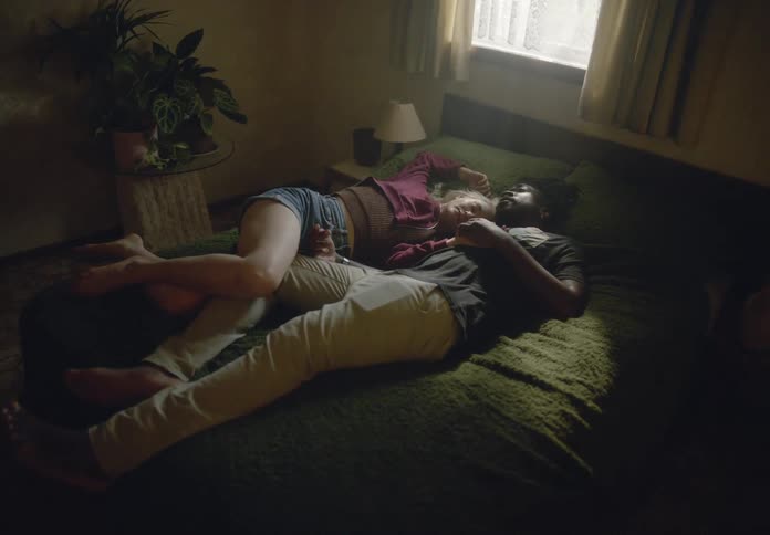 A moody intimate moment between a couple lying on a bed in a dimly lit room with a soft natural light filtering through the window