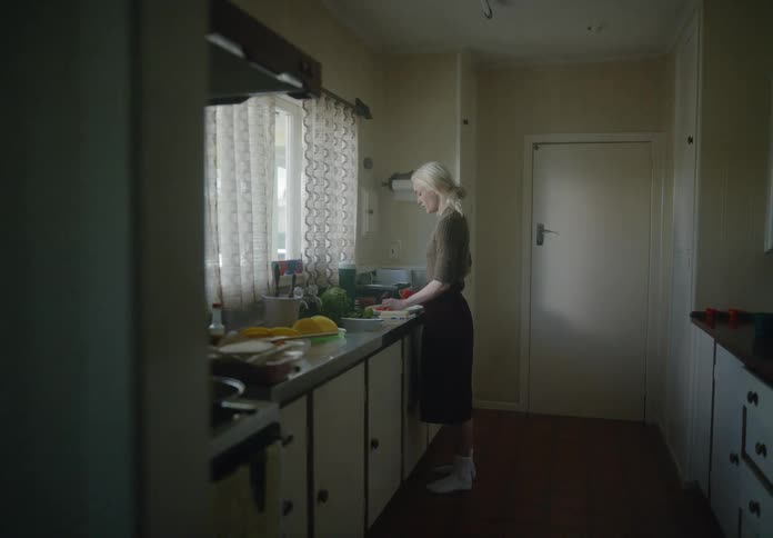 A blonde woman in a brown shirt skirt and white socks cuts vegetables in the kitchen