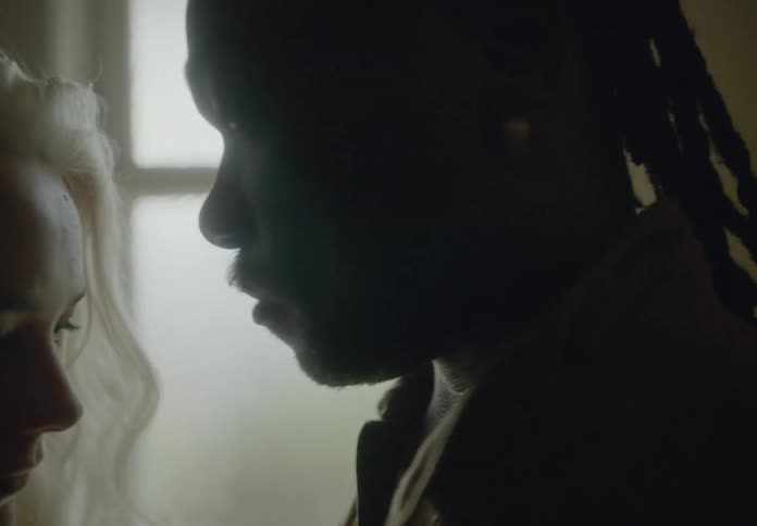 A blonde woman and a dark skinned man with dreadlocks stand face to face with an expression of intimacy