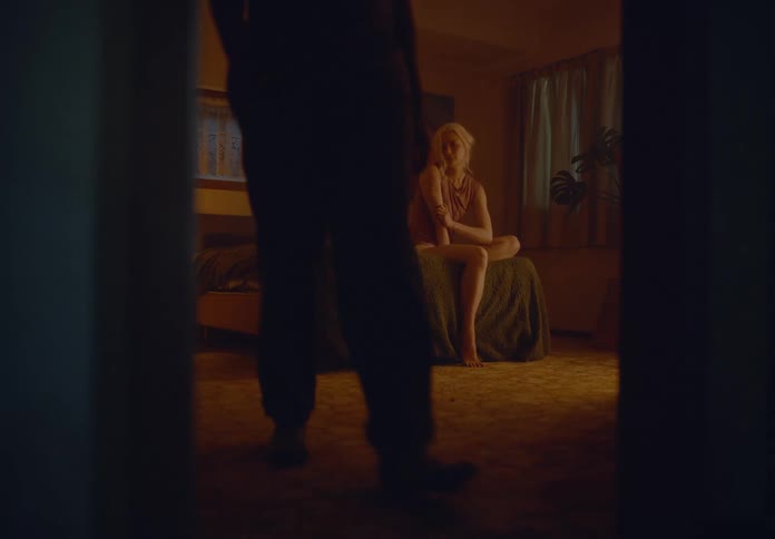 A woman sits on a bed in a dimly lit room while a man stands up and walks out conveying a sense of sadness