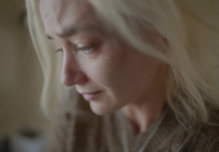 Blonde woman in a brown sweater crying near a window indoors