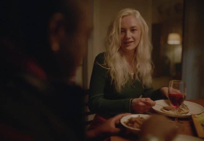 A blonde woman at a dimly lit dinner table laughs and gestures towards someone out of frame while enjoying a meal with wine