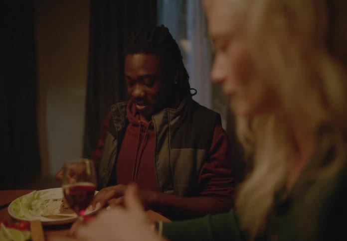 A man with dreadlocks wearing a hoodie and vest is sitting at a dimly lit dinner table smiling while cutting his food while a blonde woman in the foreground appears out of focus