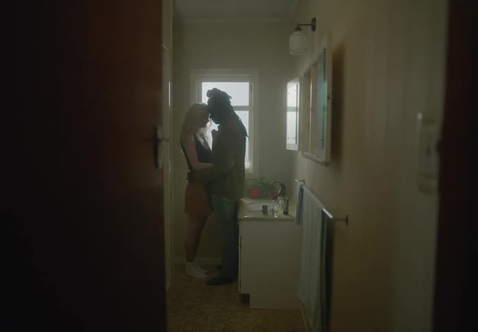 In a dimly lit bathroom a woman applying makeup at the mirror is approached from behind by a man and they embrace tenderly