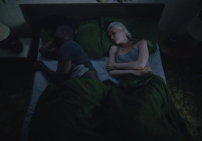 A man and a woman are lying in bed under a green blanket the man appearing upset is turned away with his arm under his head striking an attitude of frustration while the woman wearing a lightcolored tank top is awake and looking at him