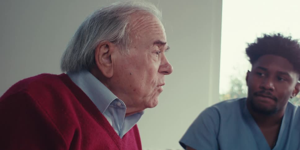 An elderly man in a red sweater talks to a healthcare worker in blue scrubs
