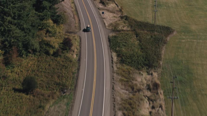 An asphalt road cuts through a rural area of woods and patchy fields