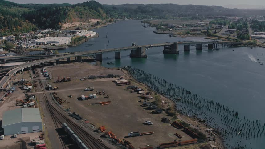 A bridge spans a wide river lined with industrial buildings railroad tracks and patches of undeveloped land under a cloudy sky