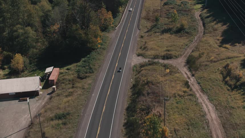 A winding road cuts through a forested landscape transitioning into autumn colors with a lone motorcycle traversing the asphalt